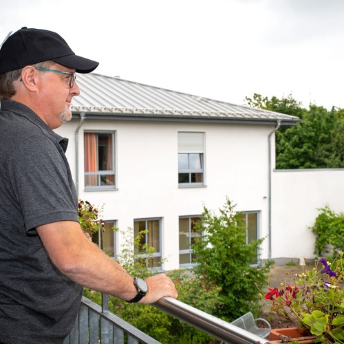 Ein Mann mit einer schwarzen Kappe steht auf einem Balkon mit Grünpflanzen und sieht in einen Garten herunter